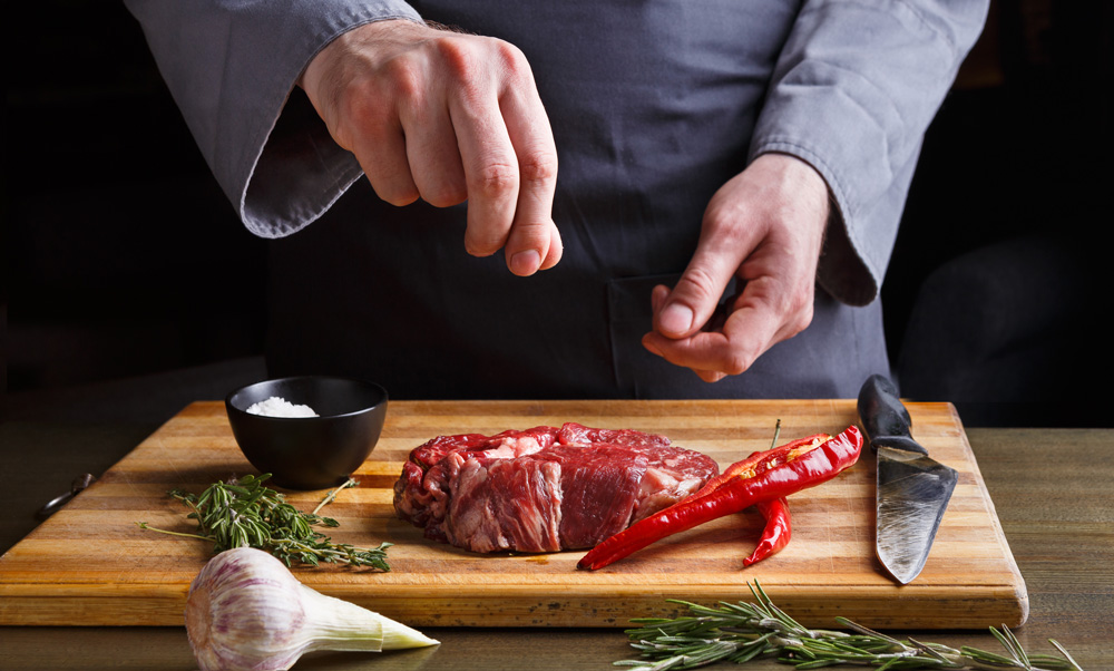 Male chef seasoning rib eye steak. Fresh meat, salt, garlic, chilli pepper and rosemary on wooden board. Modern restaurant cuisine backgroung